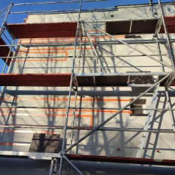 Ocieplanie elewacji budynku z użyciem styropianu, widoczne pomarańczowe paski kleju pod płyty ociepleniowe oraz rusztowanie.