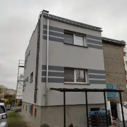 Dwu-kondygnacyjny budynek z szarą elewacją ozdobioną ciemnoszarymi pasami, widoczne rusztowanie i drabina, teren wokół zabezpieczony folią.