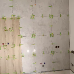 Układanie płytek ściennych w łazience z użyciem poziomowania, widoczne otwory na instalacje wodne i elektryczne, oraz fragment ściany z surowym tynkiem.