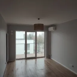 Puste, jasne pomieszczenie z parkietem, dużym oknem balkonowym, brązowym abażurem lampy i klimatyzatorem na ścianie.