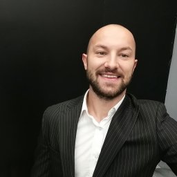 Michał Górski - Uśmiechnięty mężczyzna w garniturze w paski na tle czarnej i szarej ściany. Portret z bliska, dobre oświetlenie.