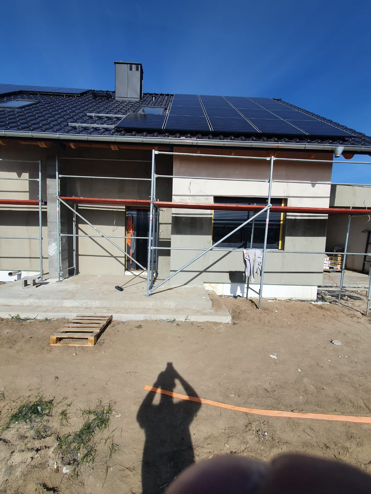 Dom w trakcie prac elewacyjnych, widoczne rusztowania, ocieplenie fundamentów, instalacja paneli słonecznych na dachu i świeżo położona elewacja w kolorze beżowym, zarys sylwetki osoby robiącej...