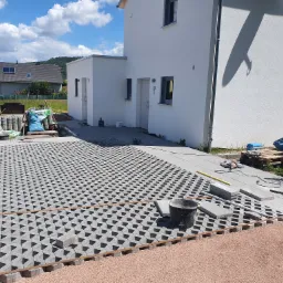 Układanie ażurowej kostki brukowej na podsypce z kruszywa przed nowoczesnym, białym domem jednorodzinnym; widoczne narzędzia, poziomica i pracownik w trakcie pracy.