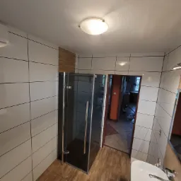 Nowoczesna łazienka z kabiną prysznicową w grafitowej ramie, białe płytki ścienne z ciemną fugą, drewnopodobna podłoga i fragment umywalki.