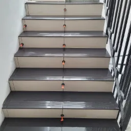 Schody w trakcie układania płytek, widoczne poziomice i krzyżyki dystansowe, górny bieg schodów jeszcze bez wykończenia, metalowa balustrada z boku.