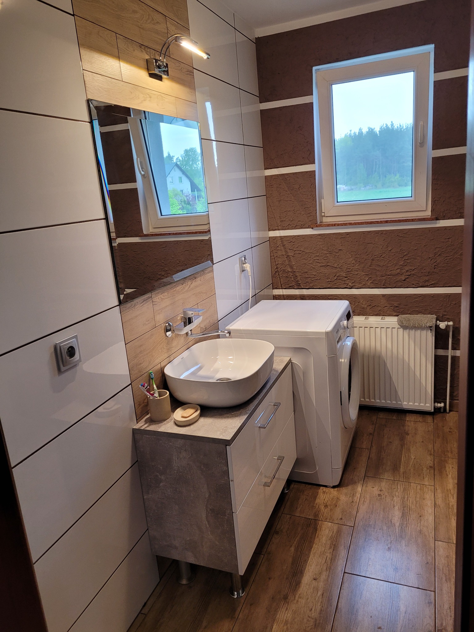 Umywalka nablatowa na szafce w nowoczesnej łazience z drewnopodobnymi płytkami na podłodze i ścianie, obok pralka i grzejnik.