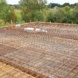 Zbrojenie płyty fundamentowej z wystającymi pionowymi prętami, widoczne rury kanalizacyjne i bloczki betonowe, w tle zielony krajobraz