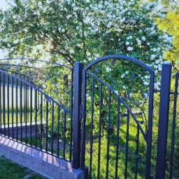 Metalowa brama wjazdowa z dekoracyjnymi elementami roślinnymi, osadzona na niskim murku z kostki, w tle kwitnące drzewo w słoneczny dzień.