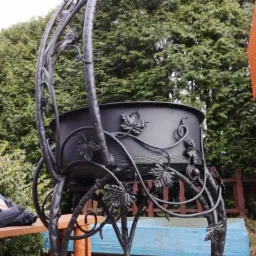 Kuty, metalowy kwietnik ogrodowy w kolorze czarnym, zdobiony motywem liści winogron, na tle zielonych drzew i drewnianego ogrodzenia.