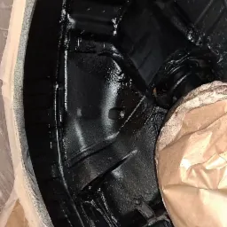 Fragment podwozia samochodu po nałożeniu czarnej, błyszczącej powłoki antykorozyjnej, zabezpieczony papierem i taśmą malarską.