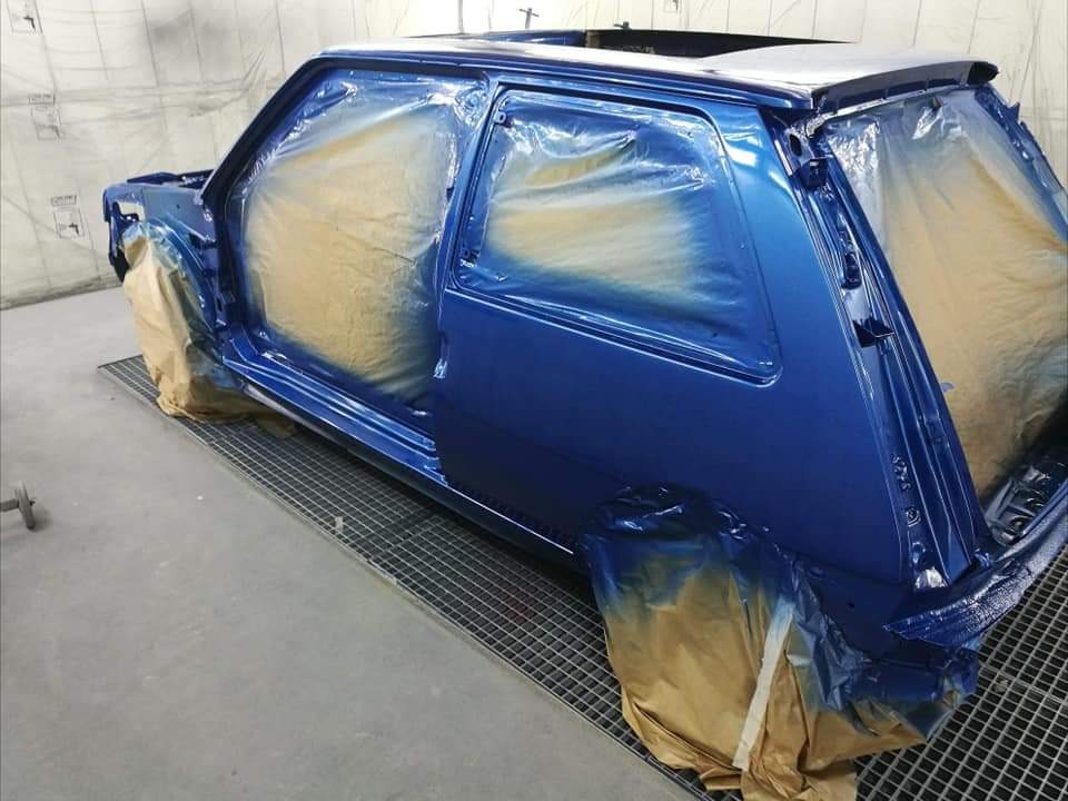 Niebieski samochód w trakcie lakierowania, zabezpieczony folią i papierem maskującym, widoczna kratka podłogowa w lakierni.