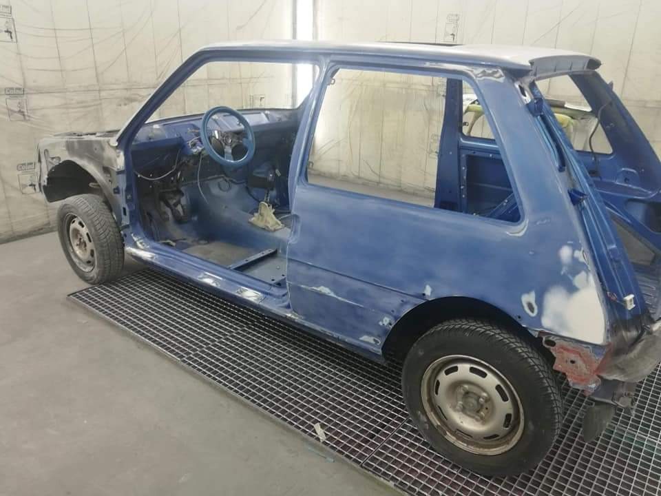 Niebieski samochód w trakcie renowacji karoserii, widoczne ślady szpachli i podkładu, wnętrze z błękitną kierownicą, na podłodze metalowa krata.