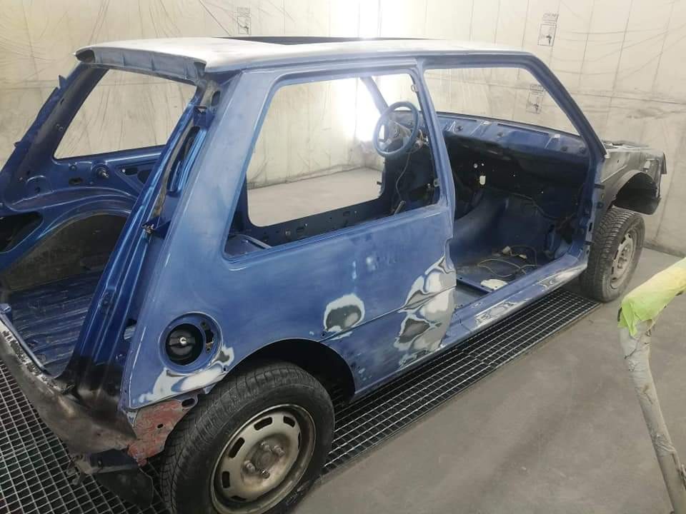 Niebieski samochód w trakcie renowacji blacharskiej, widoczne szpachlowane fragmenty karoserii i brakujące elementy, wnętrze częściowo pomalowane na niebiesko.