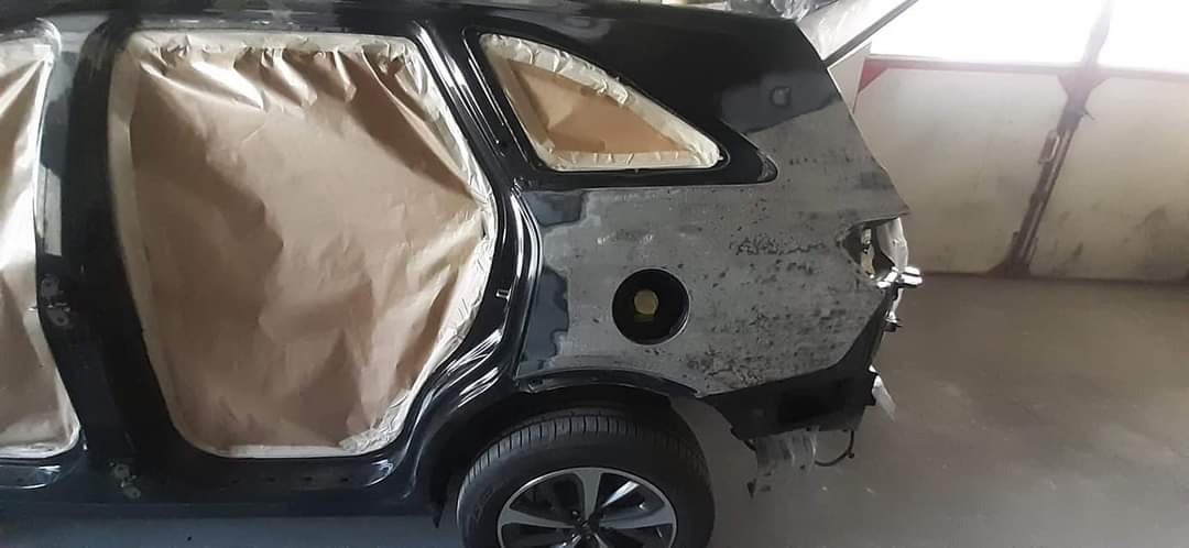 Czarne auto w trakcie naprawy blacharskiej i przygotowania do lakierowania; fragment karoserii z widocznymi śladami szpachlowania i maskowania papierem. Widoczne koło.