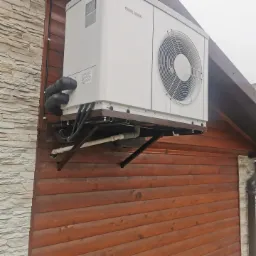 Biała pompa ciepła marki Stiebel Eltron zamontowana na brązowej, drewnianej ścianie budynku z elementami kamiennymi, widoczne rury i kable instalacyjne.