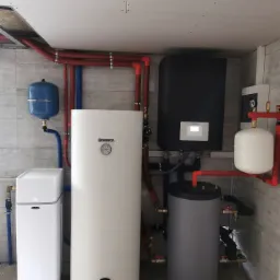 Kompletna kotłownia z różnymi urządzeniami, w tym biały zasobnik ciepłej wody marki Kospel, czarny piec z wyświetlaczem, szary zbiornik akumulacyjny i niebieskie naczynie przeponowe, połączone...