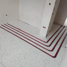 Czerwone rury ogrzewania podłogowego ułożone w pętli na podłodze w surowym stanie, widoczne ślady po instalacji w narożniku pomieszczenia.