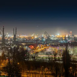 Nocny widok na portowe miasto z licznymi dźwigami, podświetlonym mostem i smugami świateł samochodowych