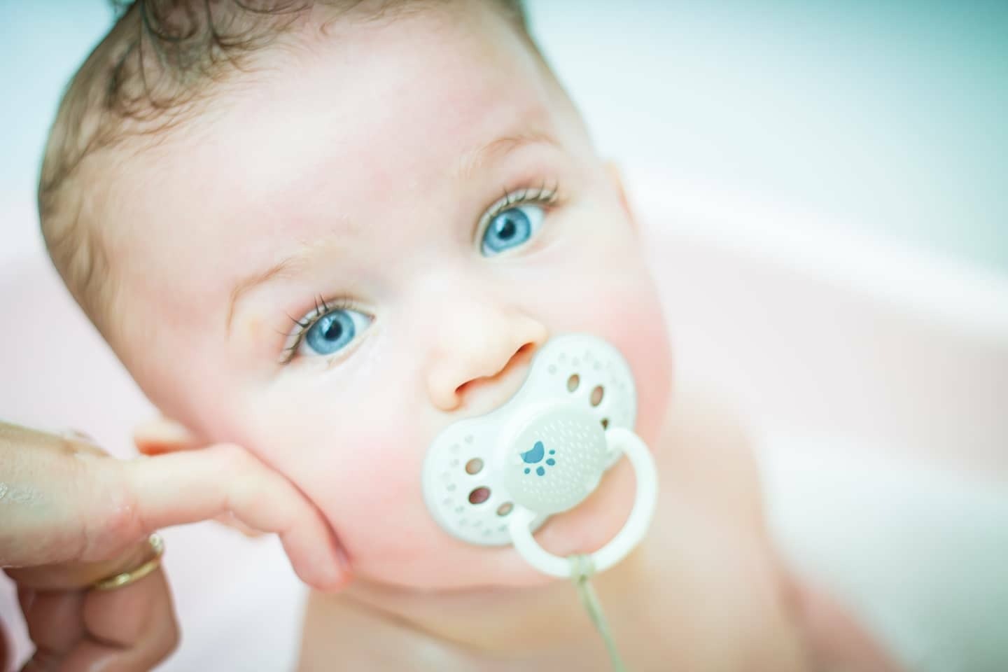 Niemowlę z jasną cerą i niebieskimi oczami, trzymające smoczek w ustach, podtrzymywane przez dłoń z pierścionkiem.