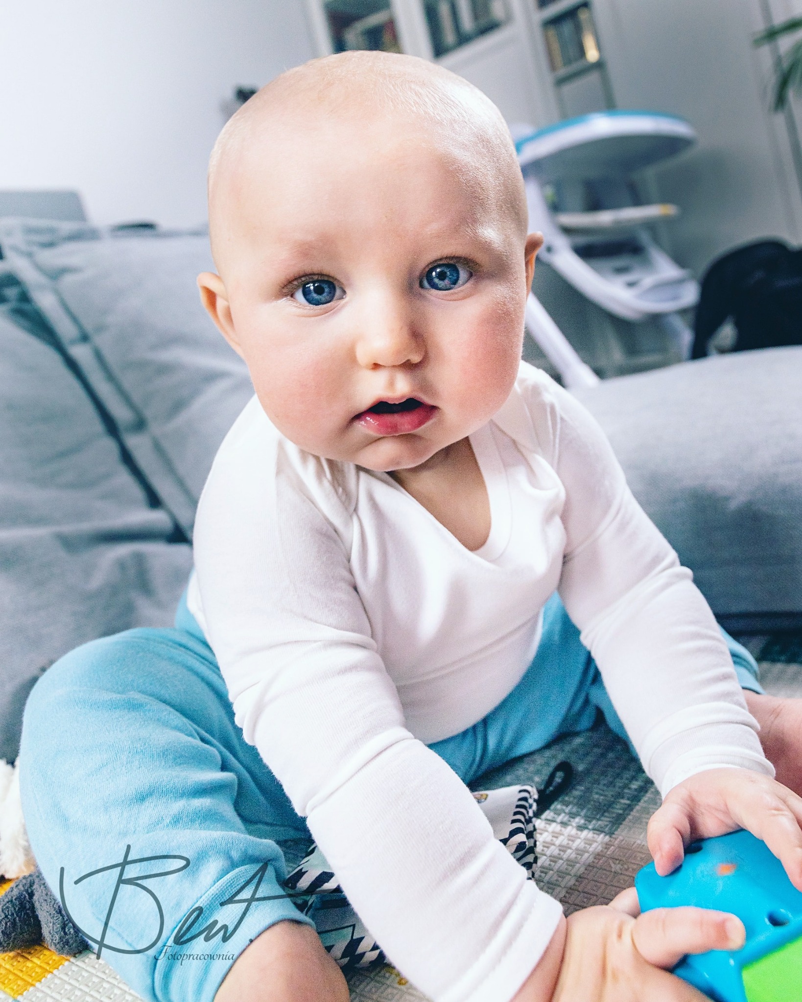 Niemowlę z dużymi niebieskimi oczami siedzi na szarych poduszkach w błękitnych spodniach i białym body, bawiąc się niebieską zabawką na wzorzystej macie.