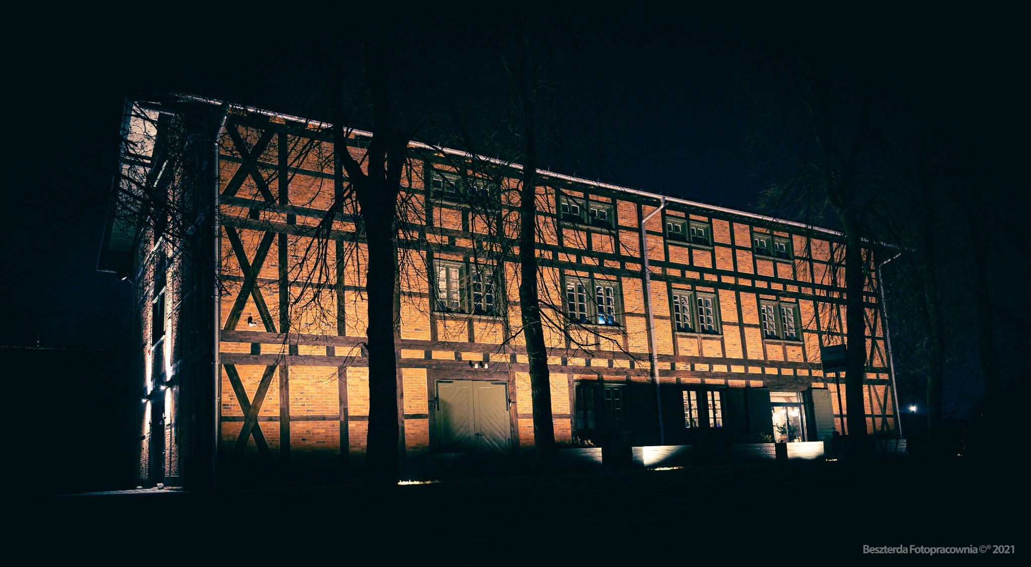 Oświetlony nocą budynek z muru pruskiego, widoczne drzewa na pierwszym planie, ciepłe światło wydobywa fakturę cegieł i drewnianych belek.