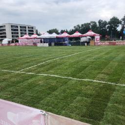 Zielona Fantazja - Rozległy trawnik z równo przystrzyżoną trawą, wyznaczony białymi liniami, w tle rozstawione namioty reklamowe z logo firmy Advantix, budynki mieszkalne i plac zabaw.