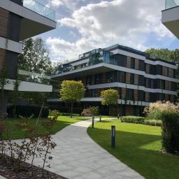 Zielona Fantazja - Nowoczesny budynek apartamentowy otoczony starannie utrzymanym trawnikiem, ścieżką z jasnoszarej kostki brukowej i niskimi drzewami, oświetlonymi przez lampy parkowe.