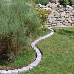 Obrzeże z kostki granitowej oddziela trawnik od rabaty z roślinnością, w tle kamienny murek.