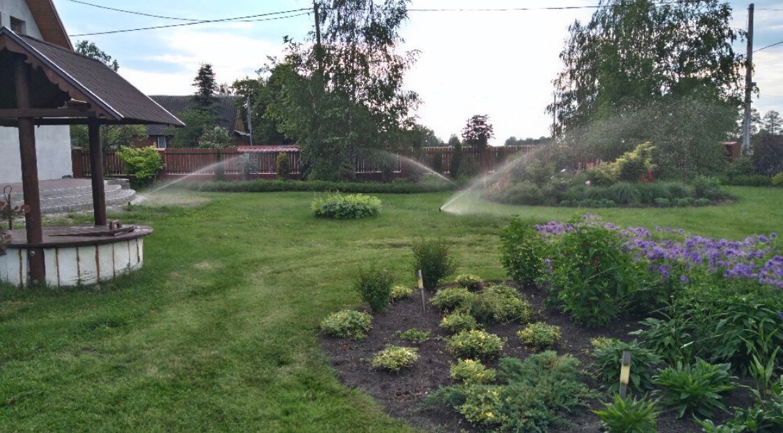 Automatyczne nawadnianie ogrodu w trakcie pracy, widoczne zraszacze rozpylające wodę na trawnik i rabaty kwiatowe, studnia z drewnianym zadaszeniem w tle.