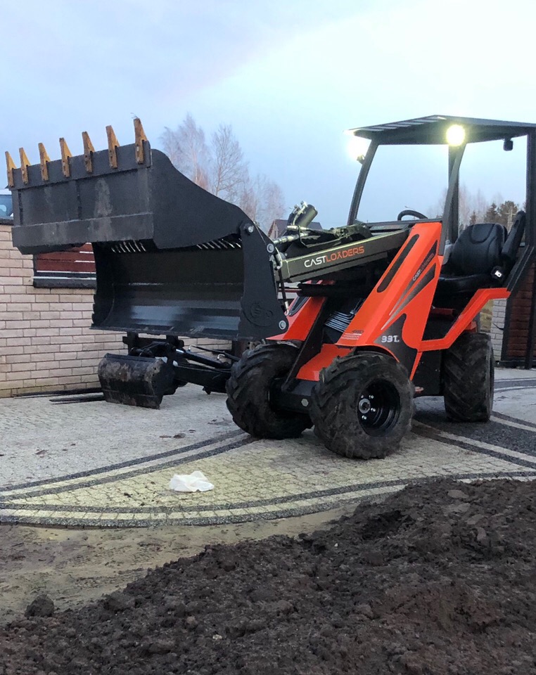 Pomarańczowa mini ładowarka Castloaders 33T z łyżką i widłami na tle kostki brukowej i pryzmy ziemi.