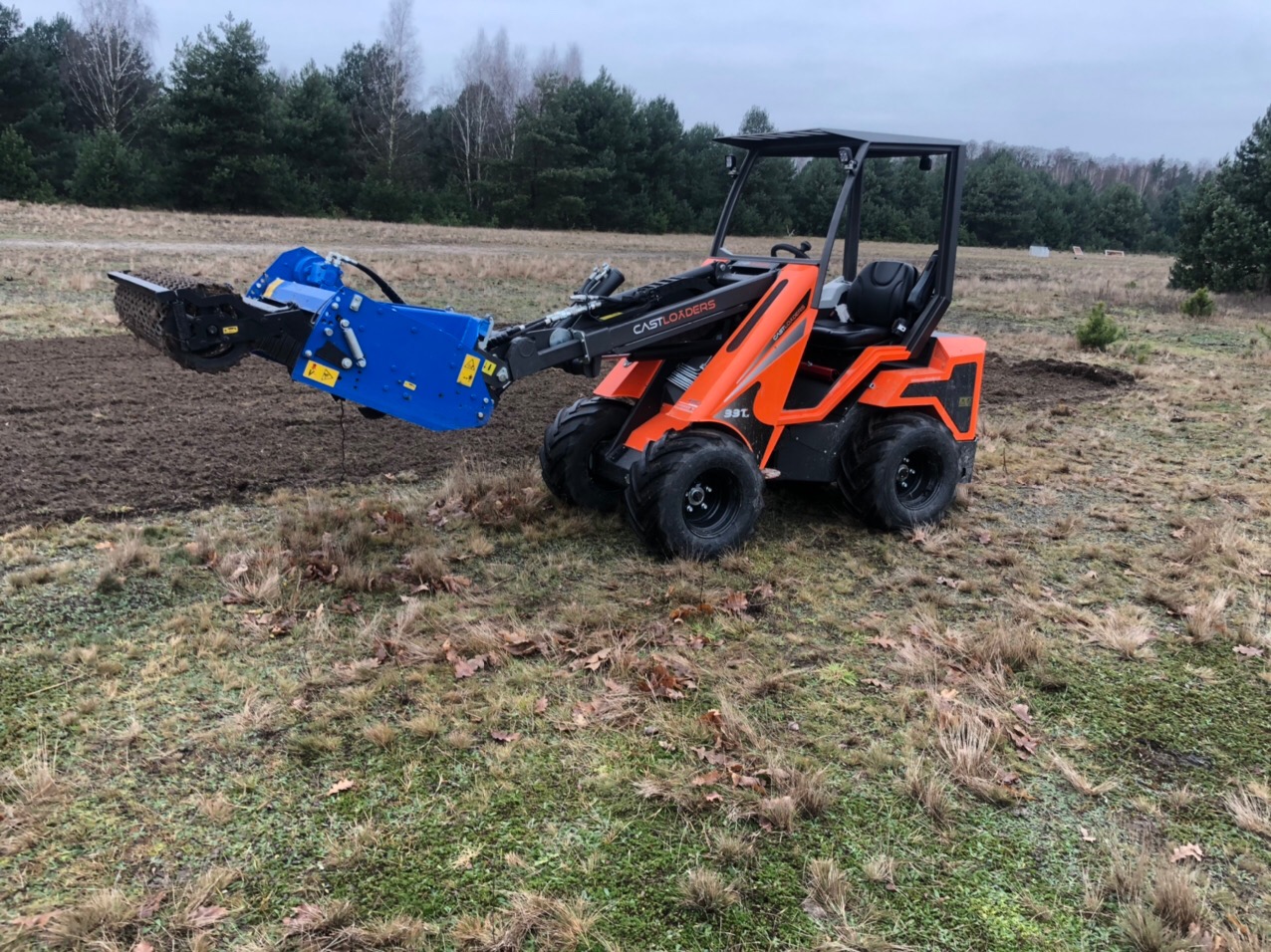 Pomarańczowa minikoparka Castloaders 33T z niebieskim osprzętem roboczym wyrównuje teren pod przyszły trawnik na tle lasu.