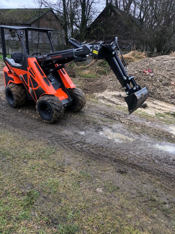 Pomarańczowa mini-ładowarka CASTLOADERS na błotnistym terenie, w tle stary budynek gospodarczy i pryzma ziemi.