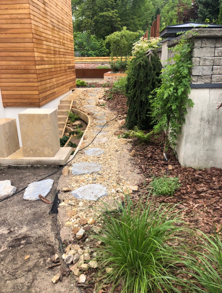 Ścieżka z kamiennych płyt w ogrodzie, częściowo utwardzona, z widocznymi elementami drewnianej elewacji budynku i roślinności ozdobnej.