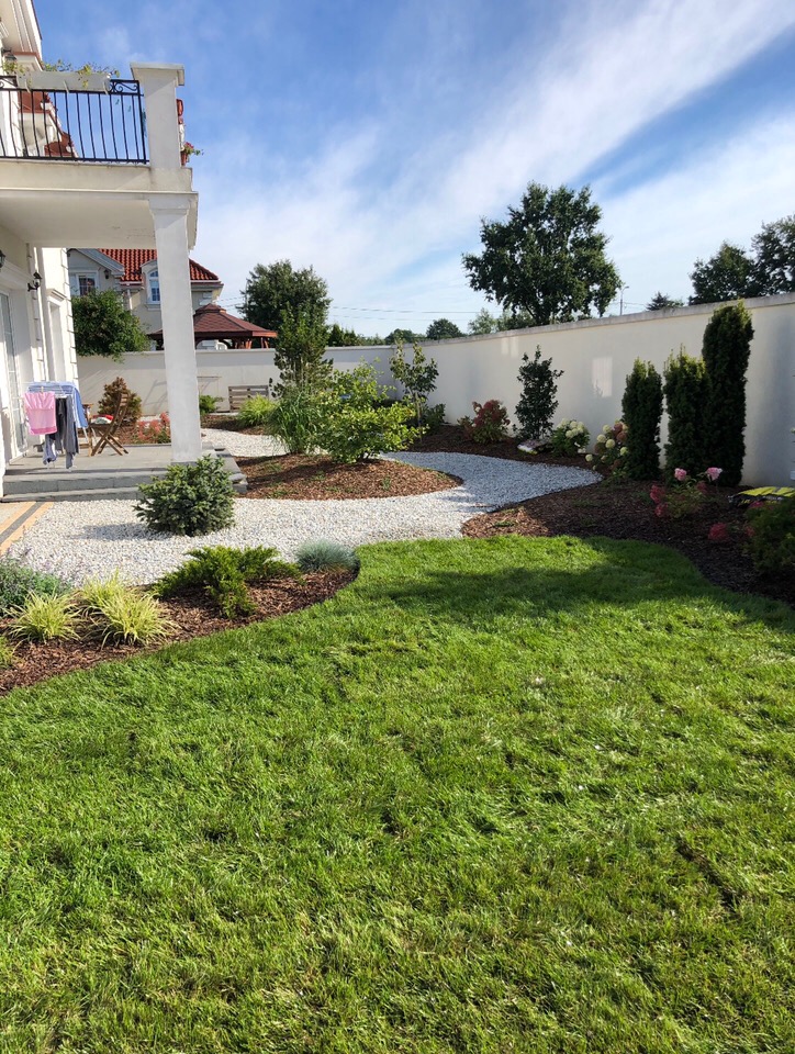 Nowoczesny ogród z białym żwirem, korą, świeżo skoszoną trawą i ozdobnymi krzewami na tle białego muru i domu z balkonem.