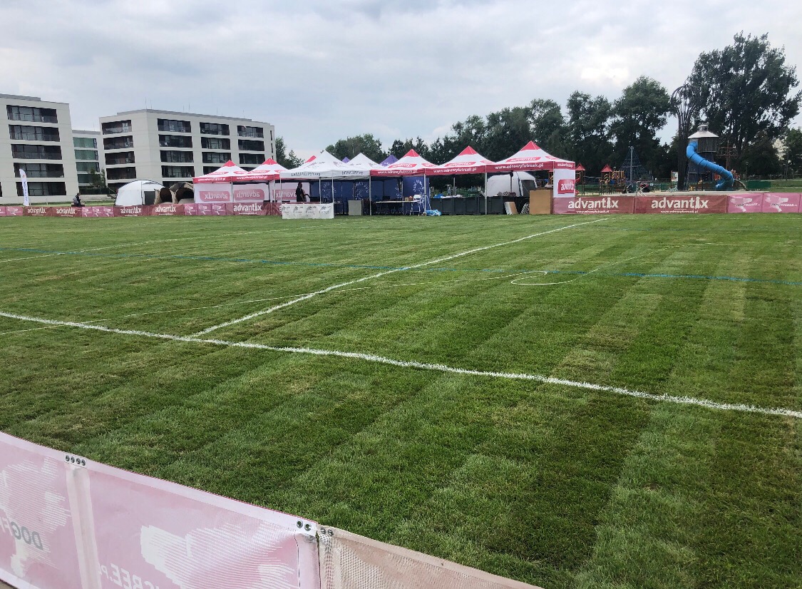 Rozległy trawnik z równo przystrzyżoną trawą, wyznaczony białymi liniami, w tle rozstawione namioty reklamowe z logo firmy Advantix, budynki mieszkalne i plac zabaw.