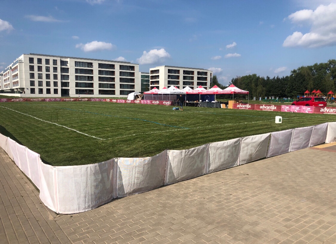 Teren zielony z równo przystrzyżoną trawą, ograniczony białym płotkiem z logo, w tle budynki mieszkalne i namioty eventowe.