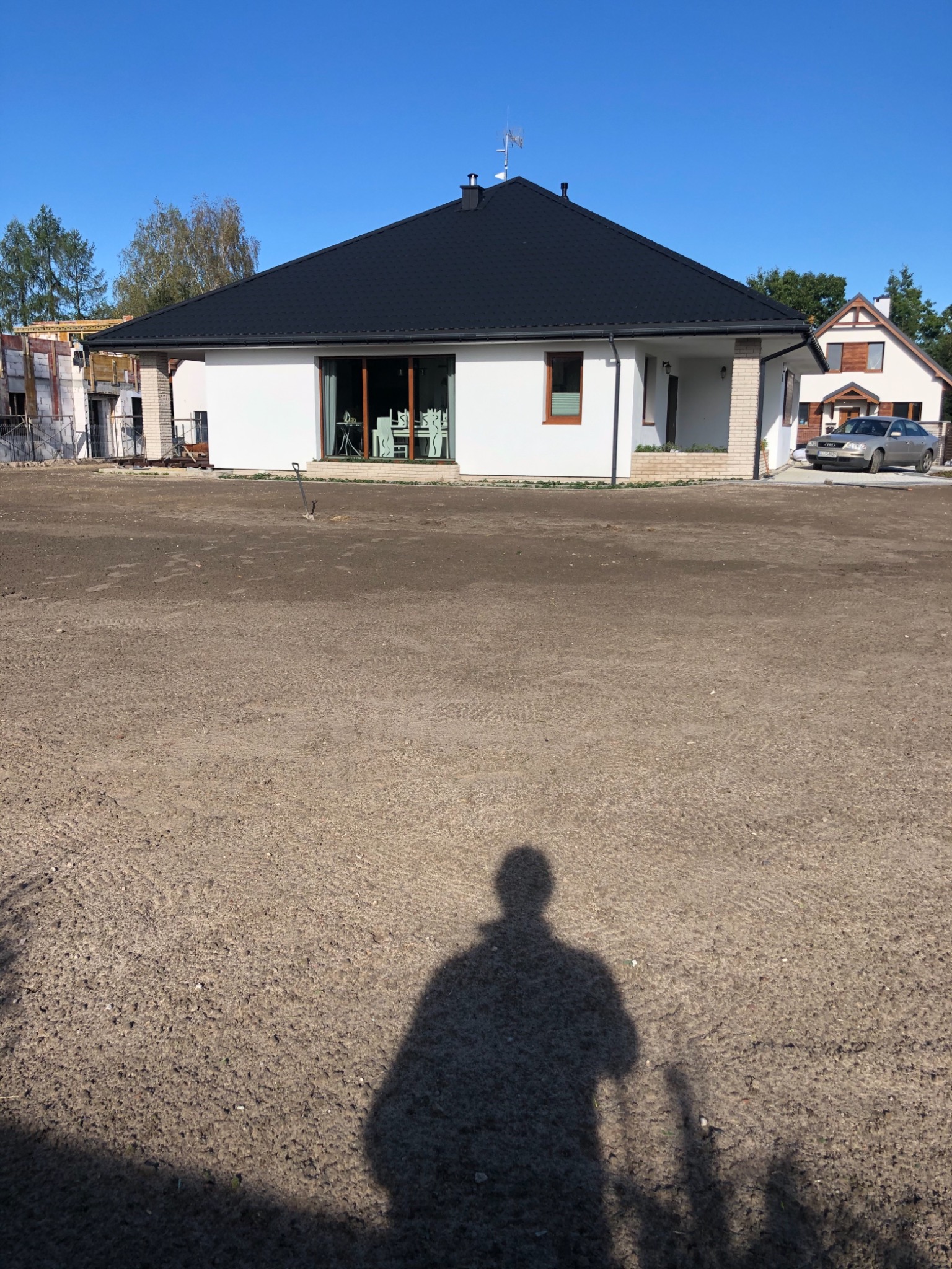 Przygotowany teren pod ogród przed nowoczesnym domem z czarnym dachem, widoczny cień osoby fotografującej i częściowo wbita szpadel.