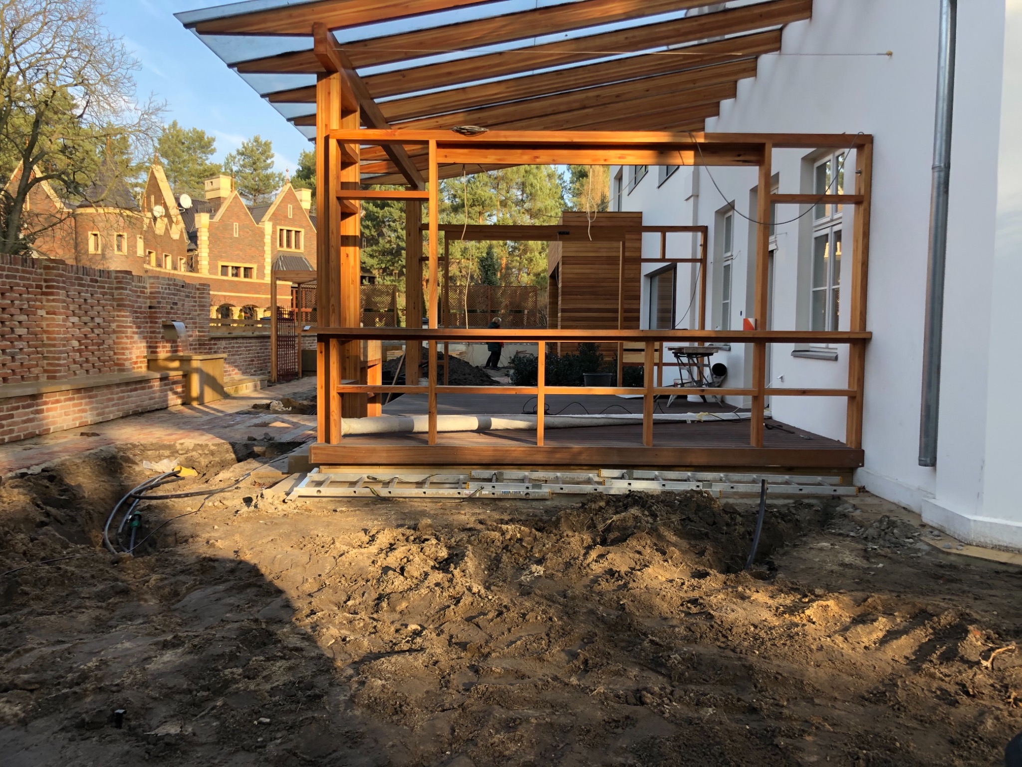 Konstrukcja drewnianego tarasu z zadaszeniem w trakcie budowy, widoczne fundamenty i instalacje w ziemi, w tle ceglany budynek.