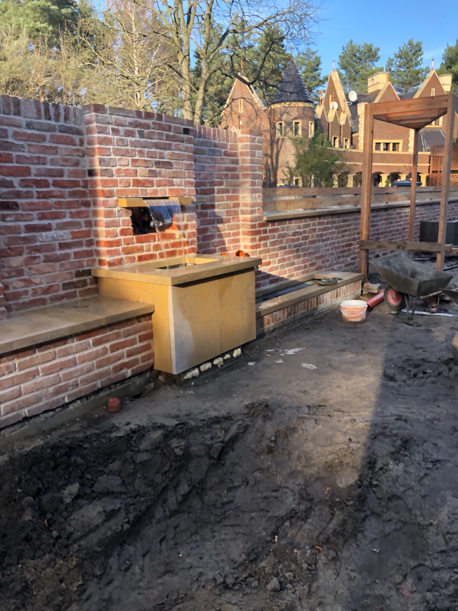 Murowana fontanna z kaskadą w trakcie budowy, widoczne narzędzia i wykop ziemny, w tle budynek z wieżyczkami.