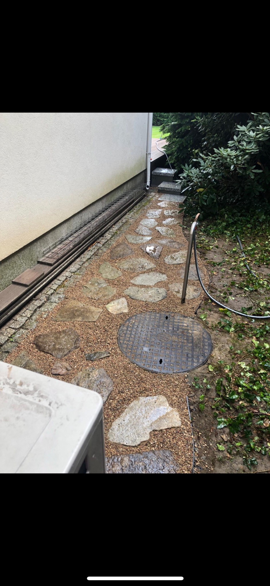Ścieżka w ogrodzie wyłożona nieregularnymi kamieniami i drobnym żwirem, z widoczną studzienką kanalizacyjną, rurą spustową i fragmentem elewacji budynku.