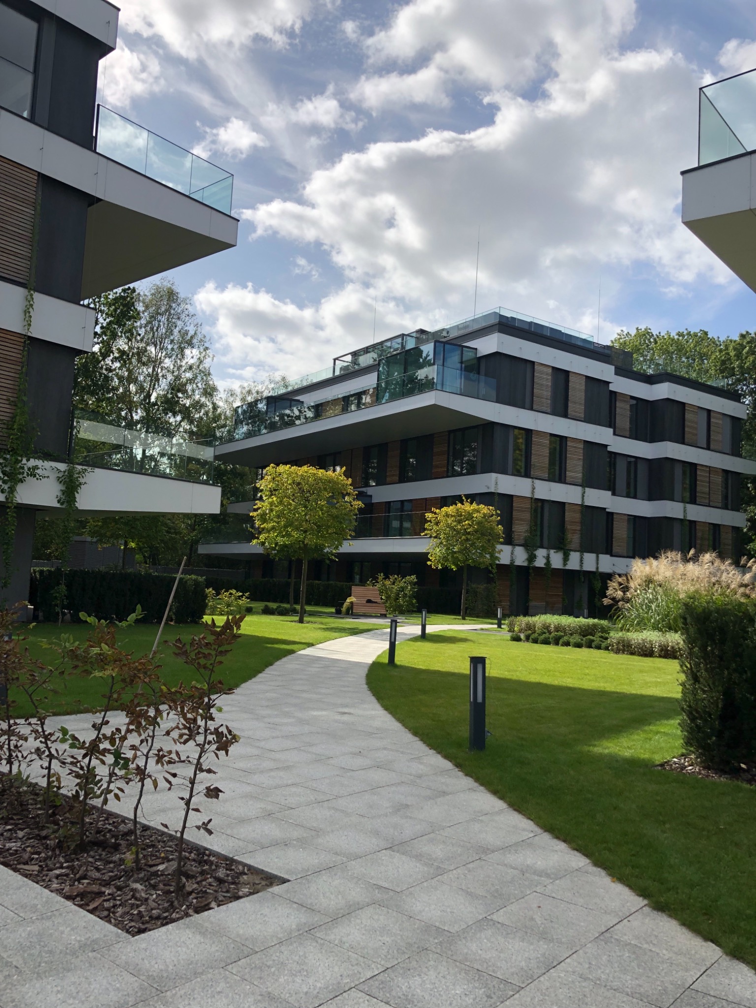 Nowoczesny budynek apartamentowy otoczony starannie utrzymanym trawnikiem, ścieżką z jasnoszarej kostki brukowej i niskimi drzewami, oświetlonymi przez lampy parkowe.