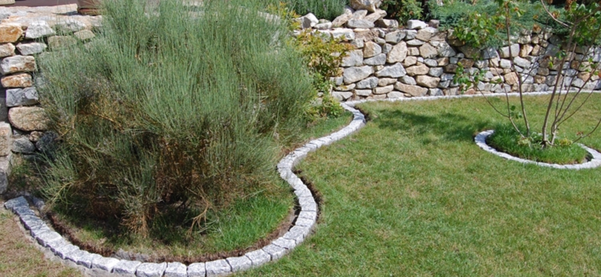 Obrzeże z kostki granitowej oddziela trawnik od rabaty z roślinnością, w tle kamienny murek.