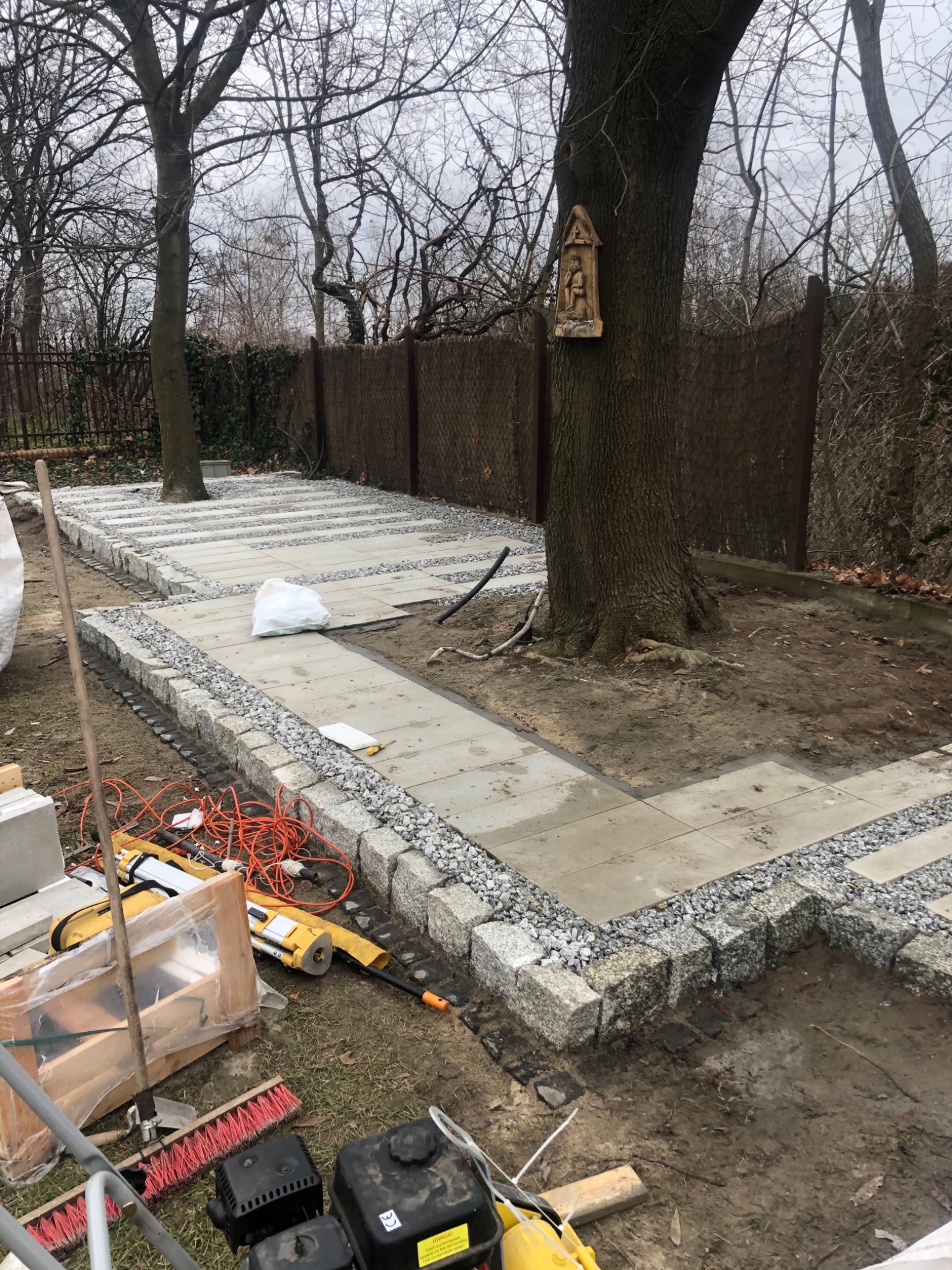 Układanie chodnika z płyt betonowych i obrzeży z kostki granitowej w ogrodzie, widoczne narzędzia i materiały budowlane, drzewo z kapliczką w tle.