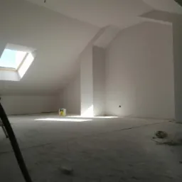 Poddasze po świeżym malowaniu na biało, widoczne okno dachowe wpuszczające światło, resztki farby na podłodze i fragment drabiny malarskiej.