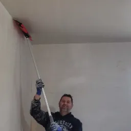Uśmiechnięty mężczyzna w roboczym ubraniu, z długim uchwytem i szpachelką w dłoniach, wygładza świeżo nałożoną masę na ścianie pod sufitem w rogu pokoju.
