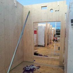 Andrzej skorupski - Wnętrze budynku w trakcie budowy z drewnianych paneli, widoczne otwory okienne i drzwiowe, metalowa listwa ukośnie oparta o ścianę, lina na podłodze, drabina w tle.