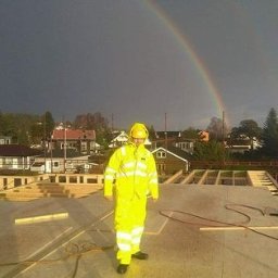 Andrzej skorupski - Pracownik w żółtym kombinezonie i kasku stoi na surowym stropie budynku w trakcie budowy, w tle tęcza na pochmurnym niebie.