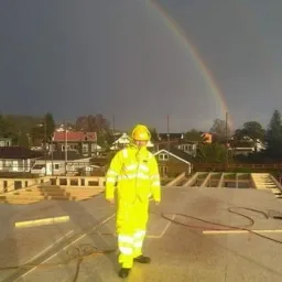 Pracownik w żółtym kombinezonie i kasku stoi na surowym stropie budynku w trakcie budowy, w tle tęcza na pochmurnym niebie.