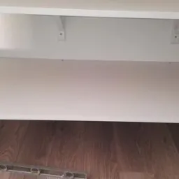 Biała zabudowa z półkami przymocowanymi do ściany, widoczny fragment brązowych paneli podłogowych i poziomica.