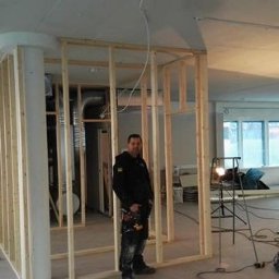 Andrzej skorupski - Konstrukcja ścian działowych z drewnianych belek w surowym stanie, widoczny pracownik z narzędziami, nieukończone wnętrze z widocznymi instalacjami i oświetleniem budowlanym.